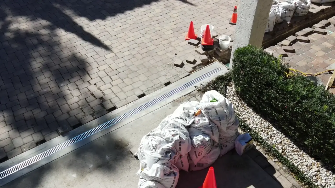 A long channel-drain grate crossing a paved area, with removed pavers, cones and material bags around the open work.